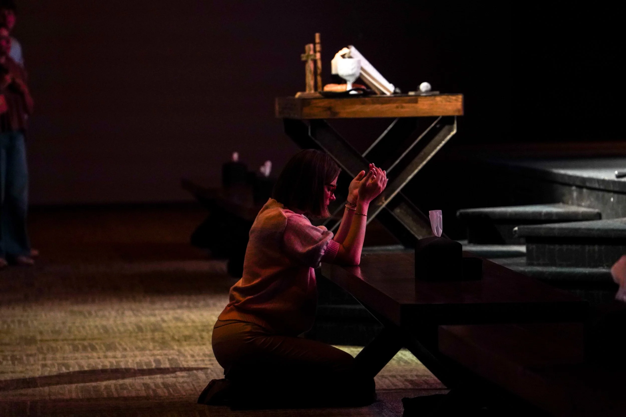 A woman kneeling and praying at a church altar during a service, with others standing in the background, dimly lit with a spotlight on her.