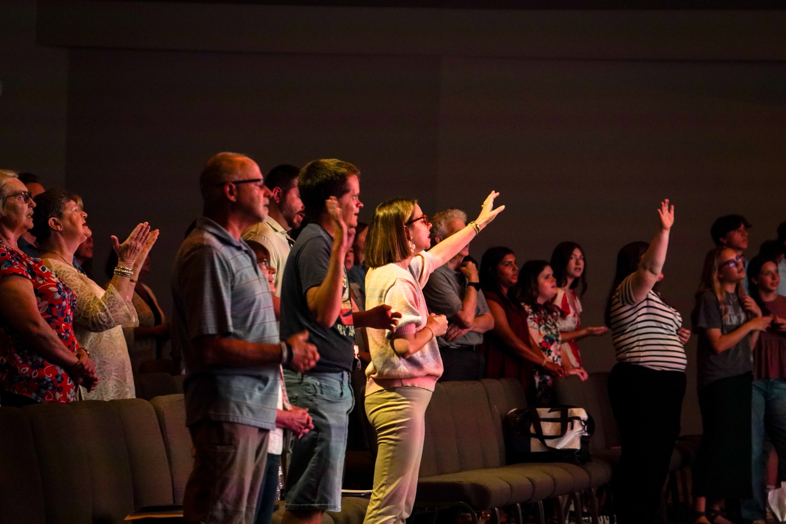 A diverse group of people standing and praying in a church or auditorium, some with eyes closed and hands raised.