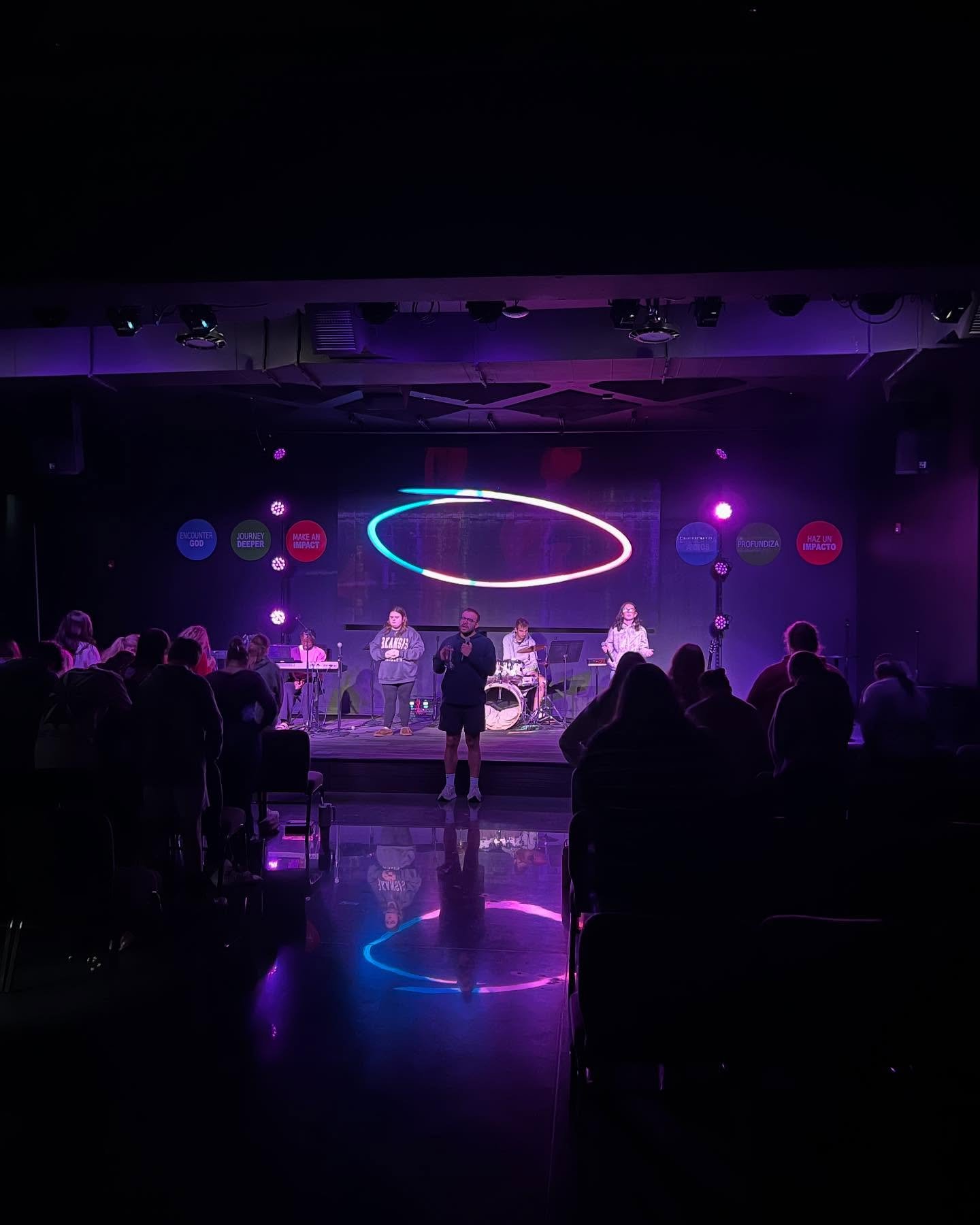 People on stage performing or speaking during a live event or concert, with colorful neon lighting and an audience watching.