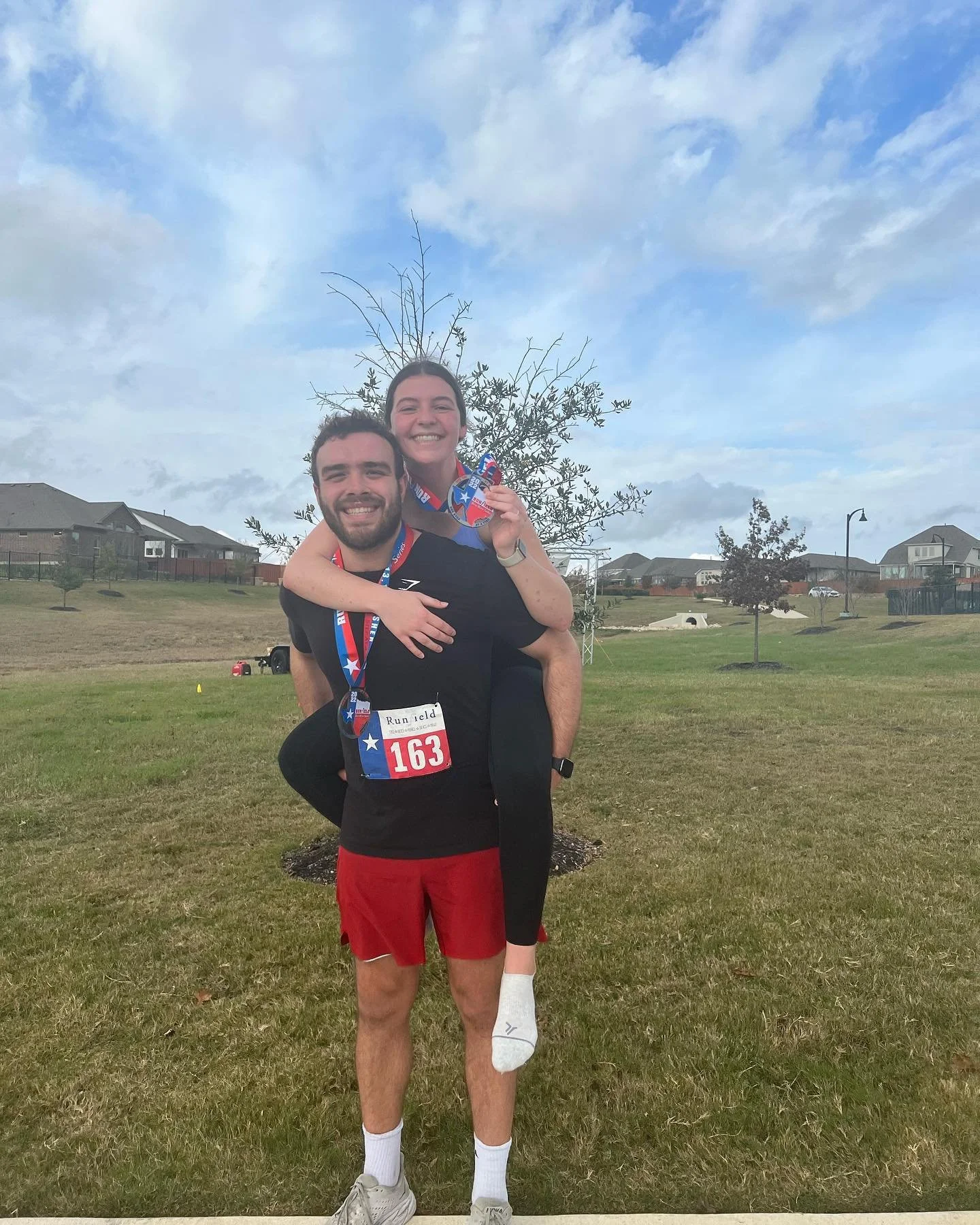 A man and woman celebrating after a race, with the woman being carried on the man's back. They are both smiling, and the woman is holding a medal. The man has a race bib number 163 and is wearing red shorts and a black T-shirt. The woman is wearing black leggings and a blue shirt. They are outdoors in a grassy area with a few trees and houses in the background, under a partly cloudy sky.