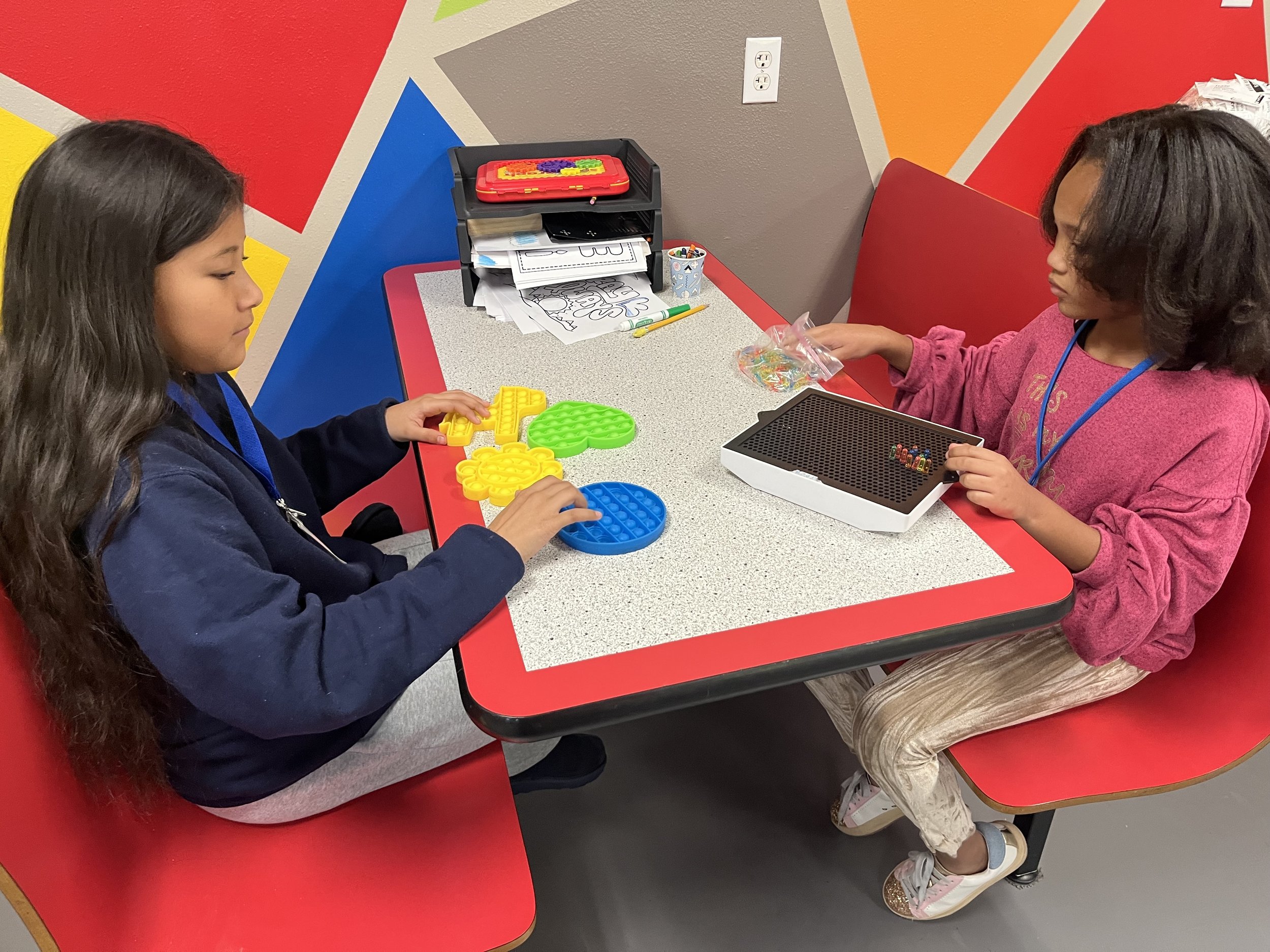 Two young girls sitting at a colorful table, playing with nerf war games, with a shelf holding papers and art supplies in the background.