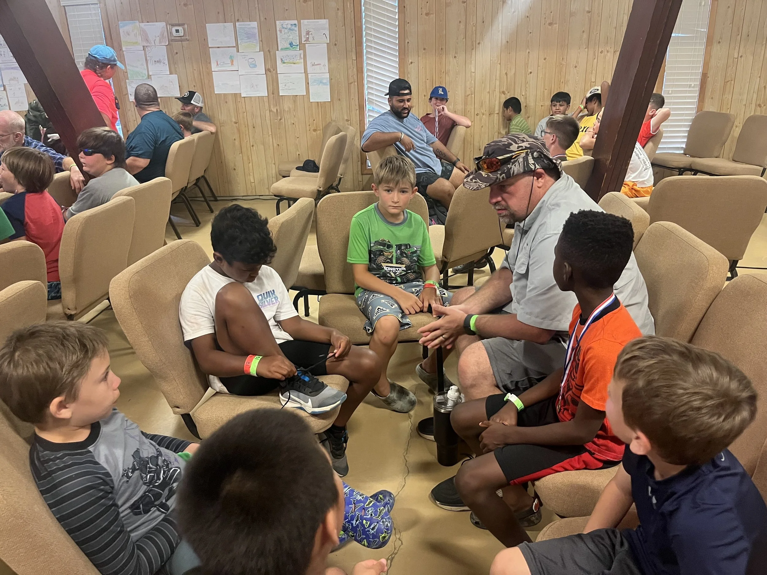 A group of children and adults sitting in a circle in a wooden room, several of them wearing colorful wristbands, engaged in conversation or listening. The room has wood-paneled walls, windows with blinds, and children's drawings on the wall.