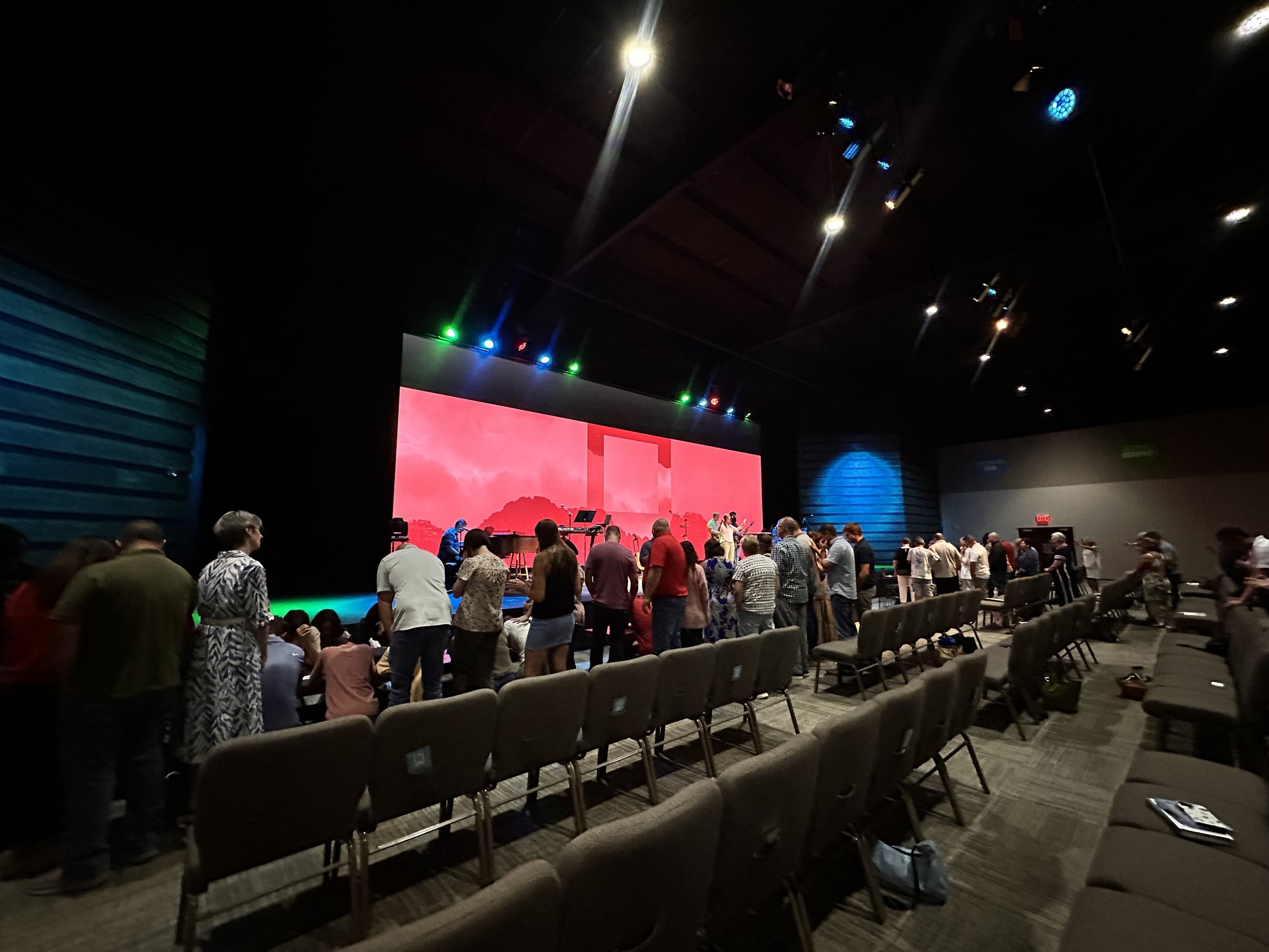 People standing and sitting in an auditorium or conference hall facing a large stage with a red digital backdrop, some musicians or performers on stage, with chairs arranged in rows and some bags on the floor.