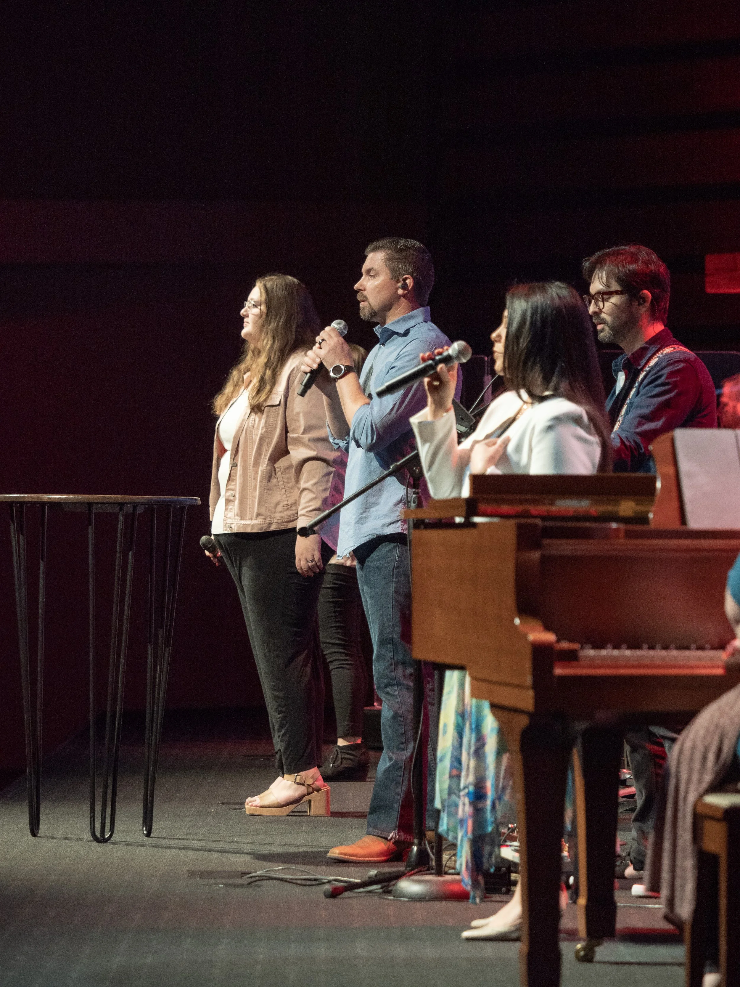 Group of five people standing on stage, holding microphones, possibly performing or speaking at an event, with a piano in the foreground.