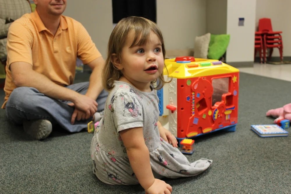 A young girl sitting on the carpeted floor in a playroom, looking surprised, with a man and children's toys around her.