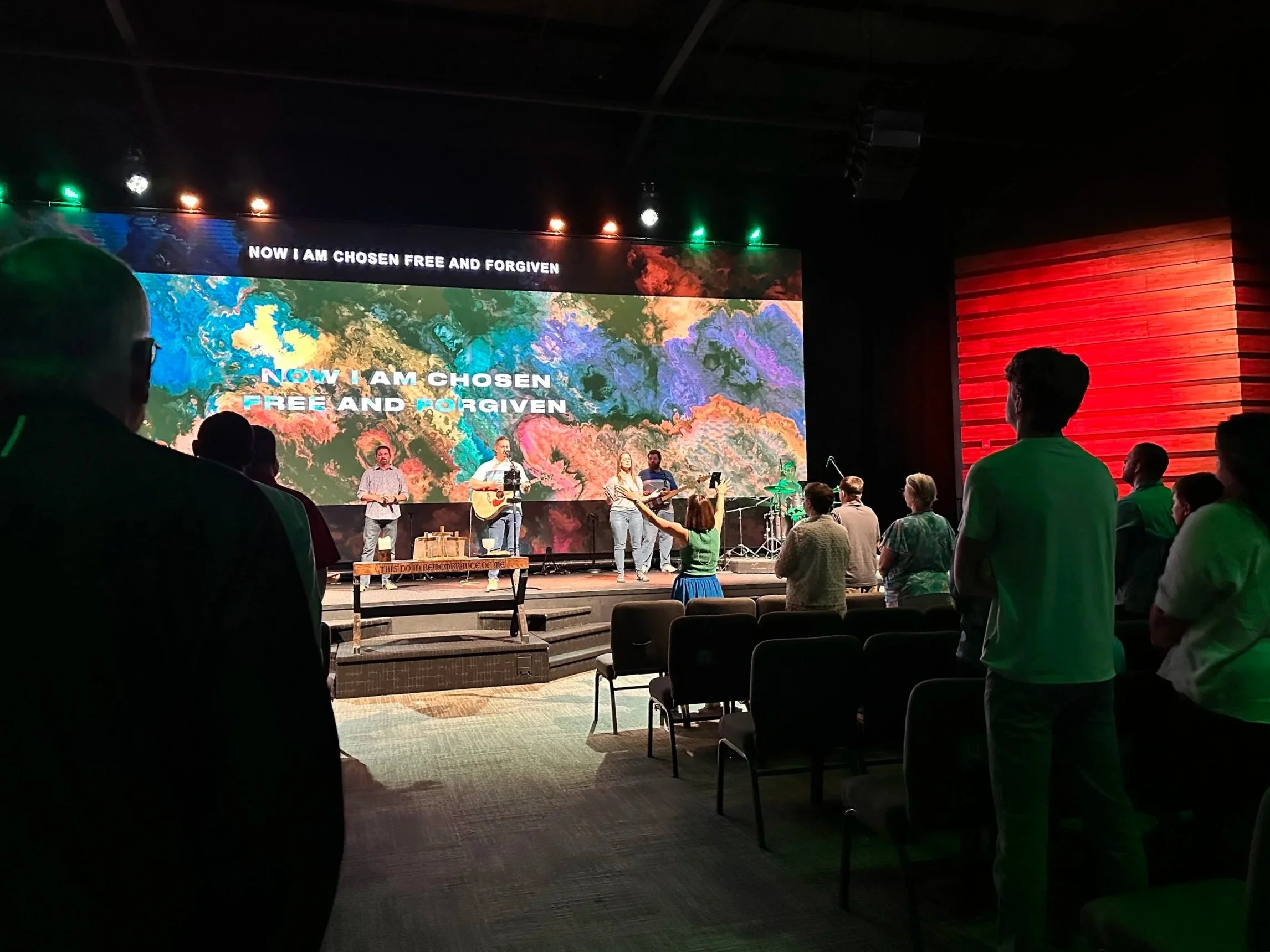 People attending a church service or concert in a dark room, facing a stage with a large screen displaying song lyrics 'Now I am chosen, free and forgiven.' Some individuals are standing, some are seated, and a band is performing on stage.