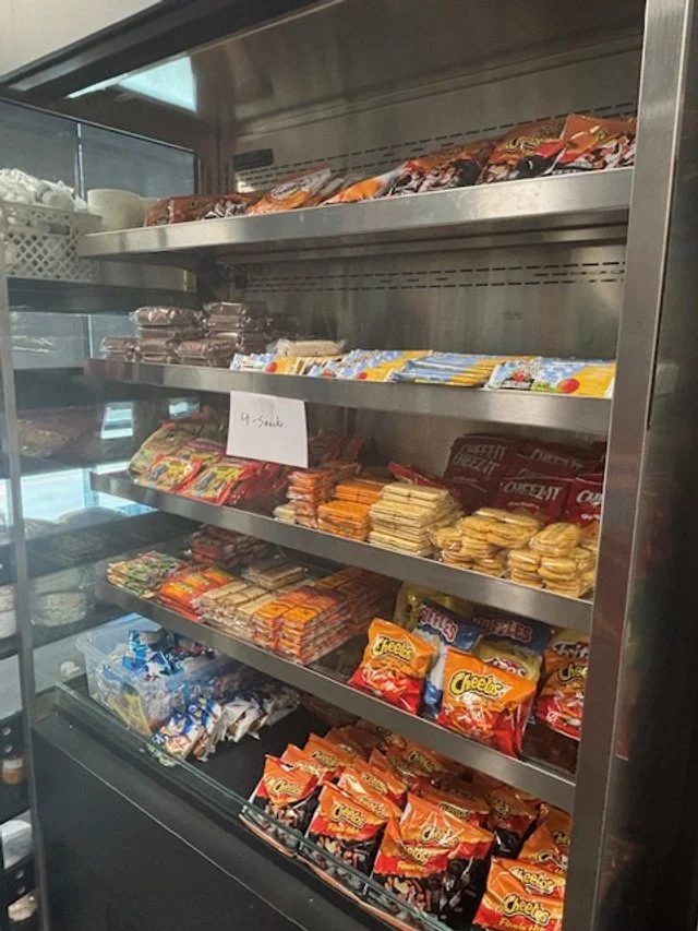 Shelves stocked with various snack foods, including Cheez-It crackers, cookies, and other packaged treats in a vending machine or snack fridge.