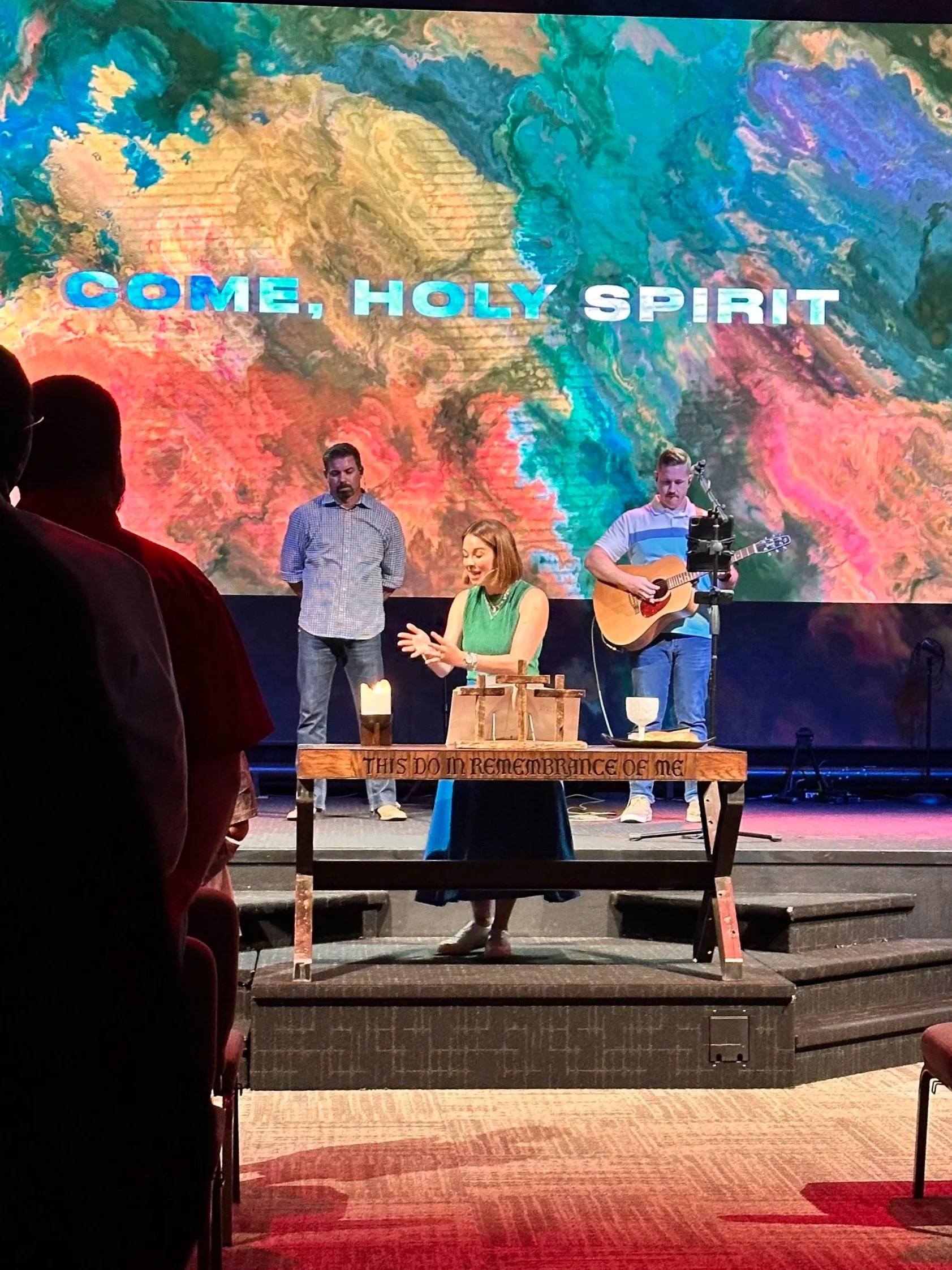 A woman on stage leading a worship service with two men behind her, one playing a guitar, in front of a large colorful backdrop with the words "COME, HOLY SPIRIT" displayed. The stage has a table with candles and a wooden cross, with an inscription that reads "THIS DO IN REMEMBRANCE OF ME." Audience members are visible in the foreground.