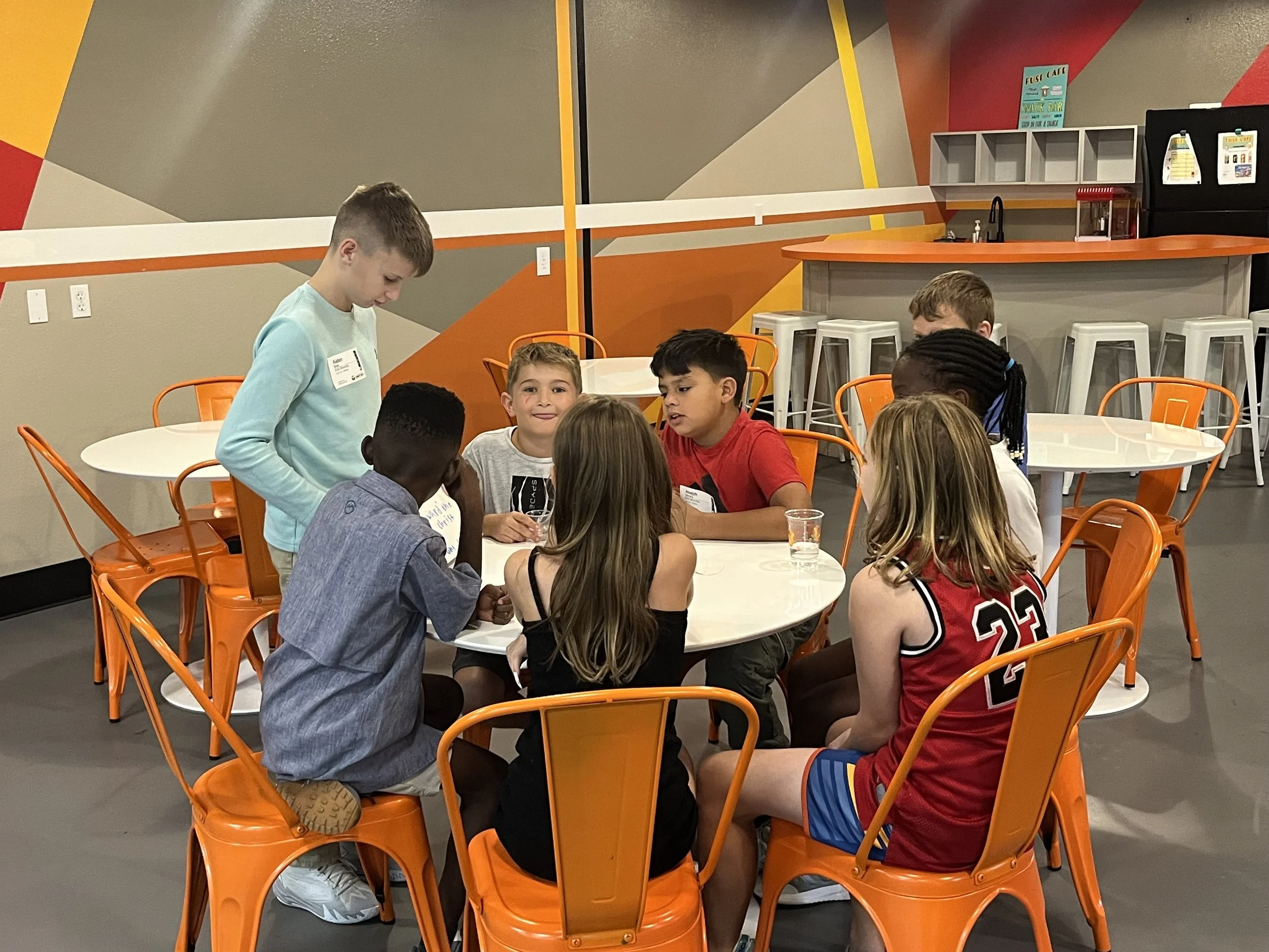 Six children gathered around a white round table in a colorful indoor space, some engaged in conversation and others looking at items on the table, with an orange and gray background.