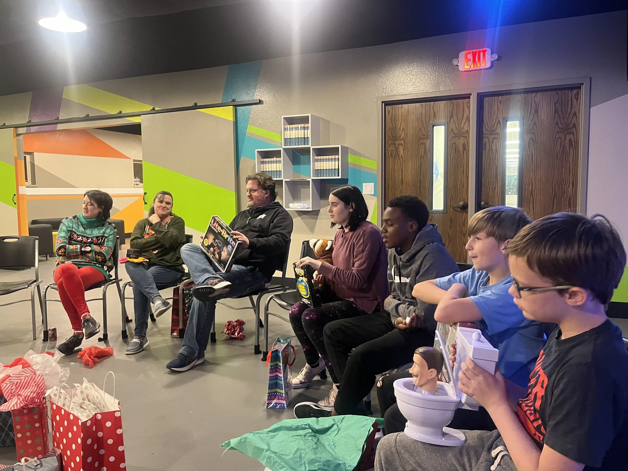 Group of people sitting in a circle during a gift exchange at a party, with presents and wrapping paper on the floor.