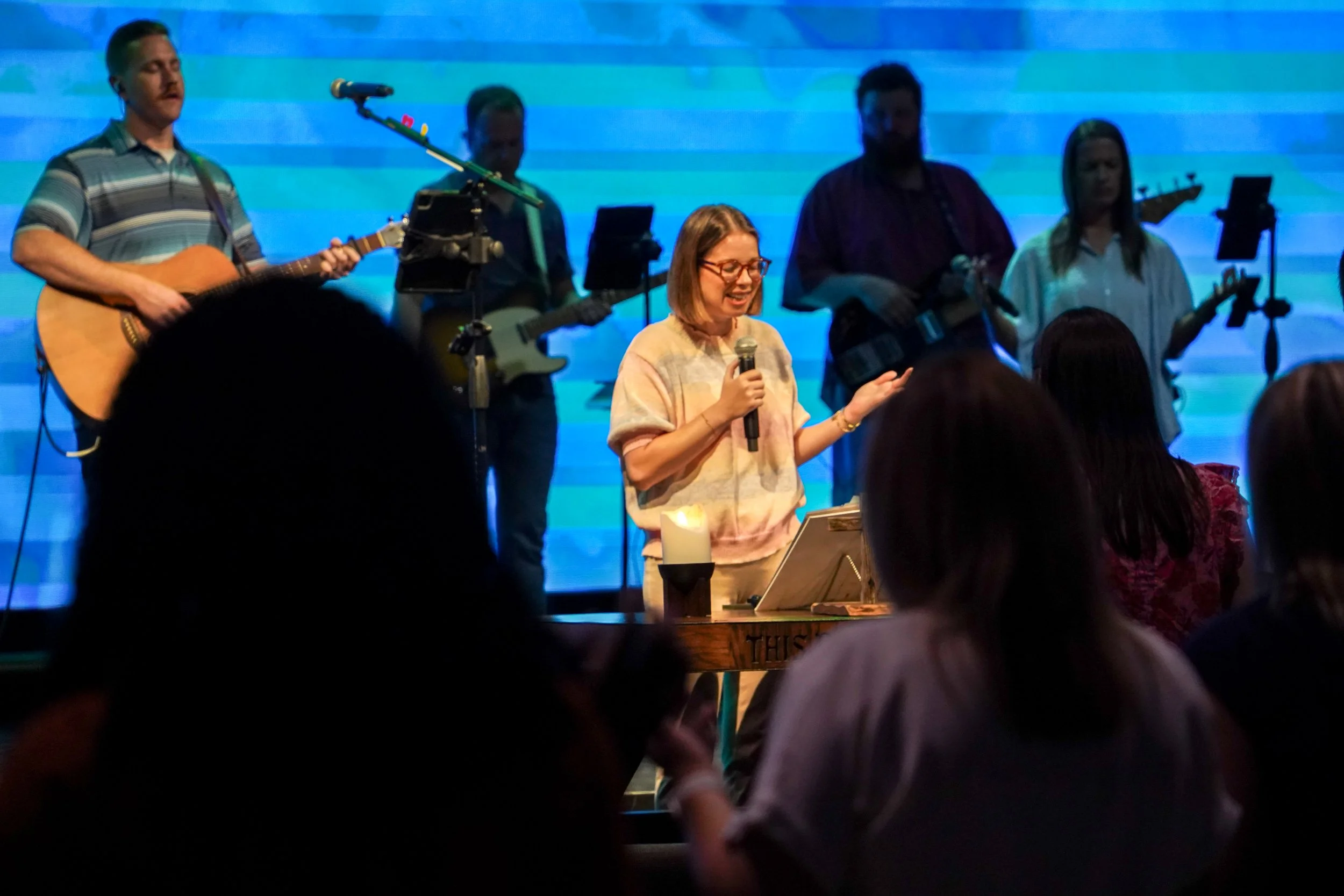 Woman with glasses holding a microphone and smiling in front of a band with guitars, while audience members watch.