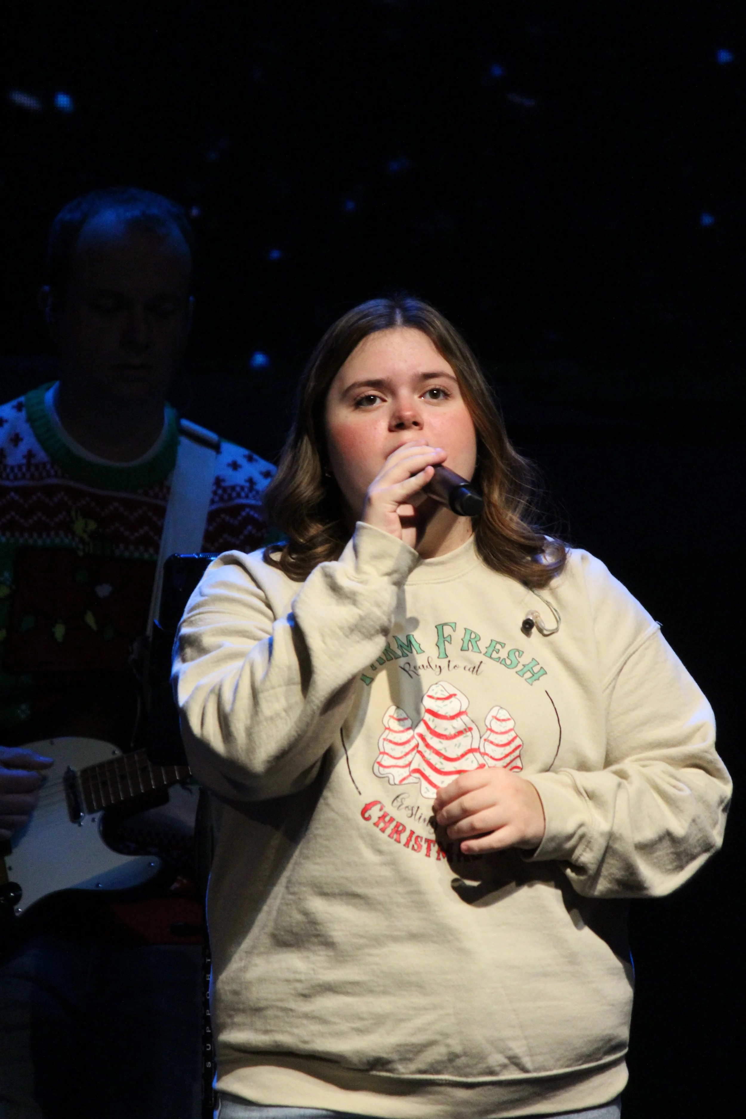 A young woman singing into a microphone on stage with a man playing guitar in the background, both under a starry backdrop.