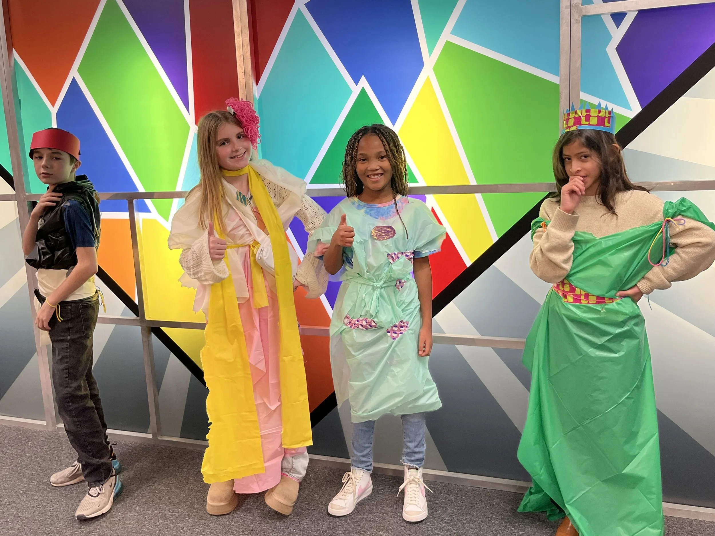 Four children dressed in colorful, creative outfits standing in front of a geometric, rainbow-colored mural. They are smiling and posing for the photo, with two giving thumbs up and one holding her chin thoughtfully.