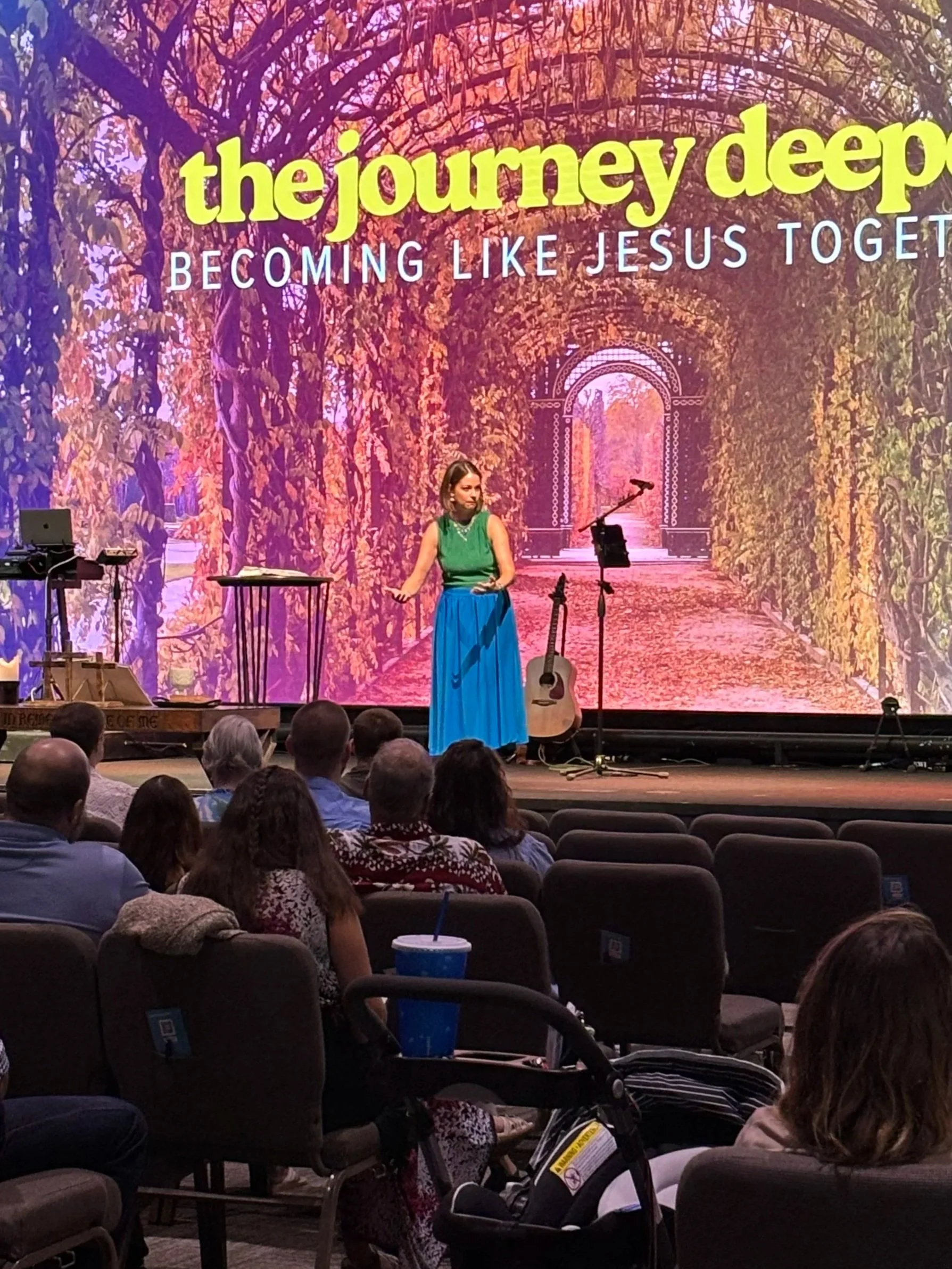 A woman on a stage giving a presentation with a large screen behind her displaying the text 'the journey deep becoming like Jesus together' over a background of a tunnel of trees with autumn leaves. There are musical instruments on the stage and an audience seated in front.