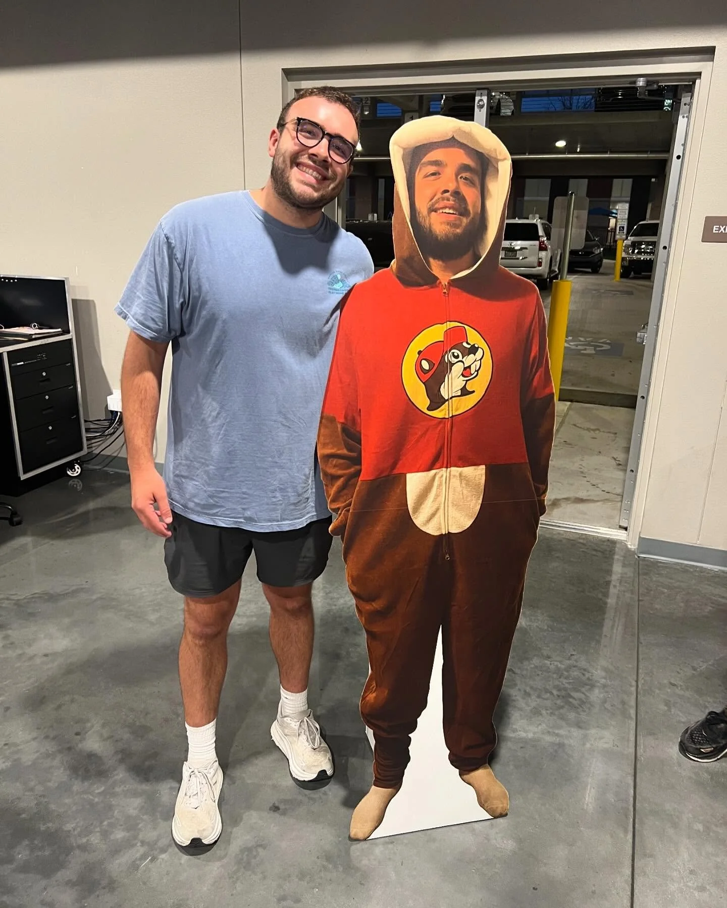 A man smiling and standing next to a cardboard cutout of himself in a costume, which is designed like a hoodie with a cartoon dog logo. The man is wearing glasses, a light blue t-shirt, and dark shorts, inside a building near an open garage door where cars are parked outside.