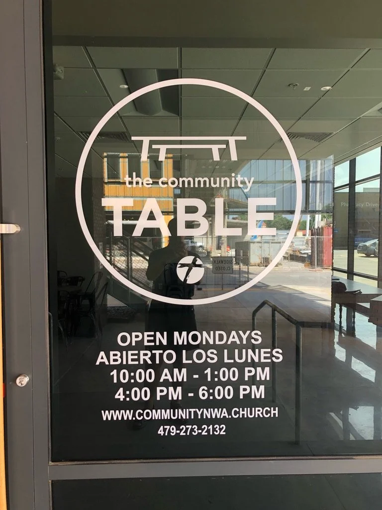 Glass door with church logo and text indicating the community Table open Mondays, bilingual in English and Spanish, with service hours, website, and phone number.