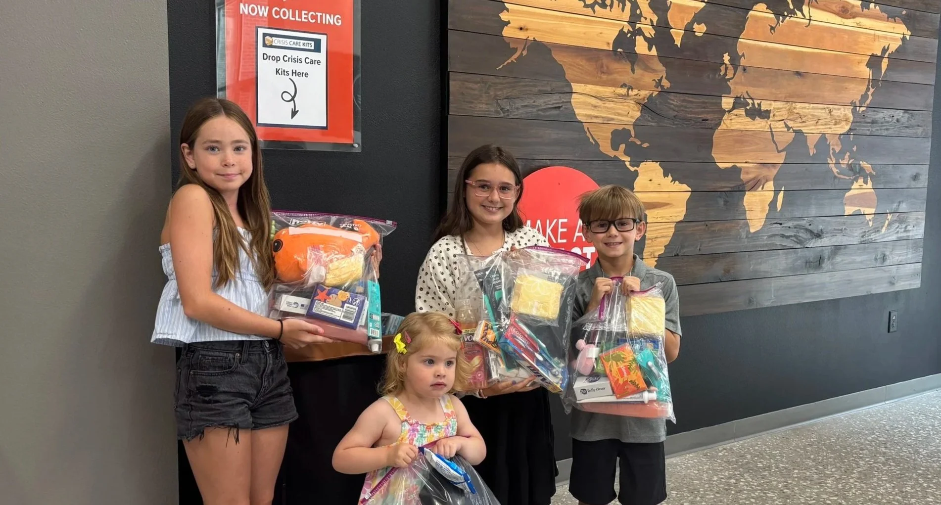 Four children standing indoors, holding plastic bags filled with various items, near a wall with a world map and a sign about collecting crisis care kits.