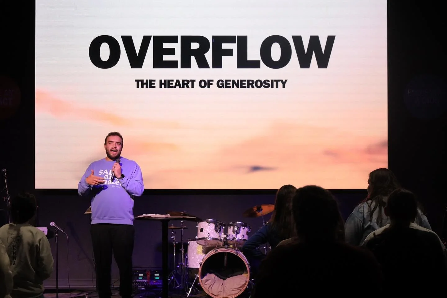 A man with a beard and light purple sweatshirt speaking into a microphone on stage in front of a large screen that reads "Overflow The Heart of Generosity." There are musical instruments and an audience in the foreground.