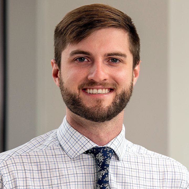 A young man with brown hair and a beard, smiling, wearing a white checkered shirt and a navy blue patterned tie, standing indoors with a neutral background.