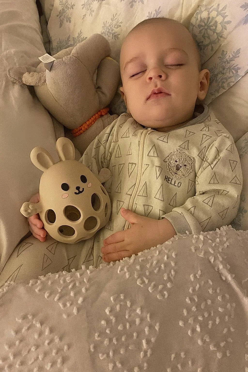 A young child is sleeping peacefully in bed, holding a beige toy with animal features and cut-out holes, surrounded by soft pillows and blankets.
