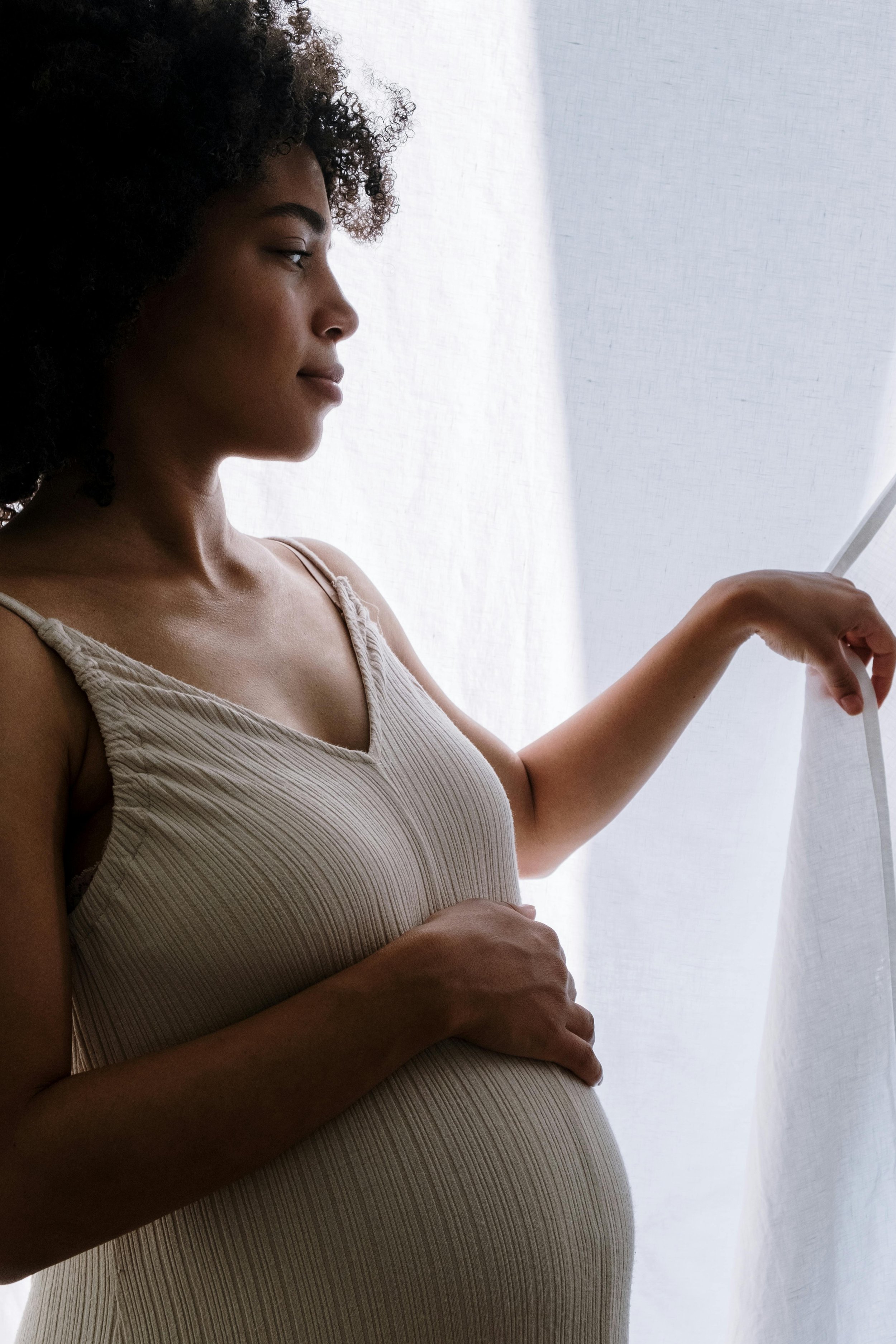 A pregnant woman standing near a white curtain, gazing out, with her hand resting on her belly.