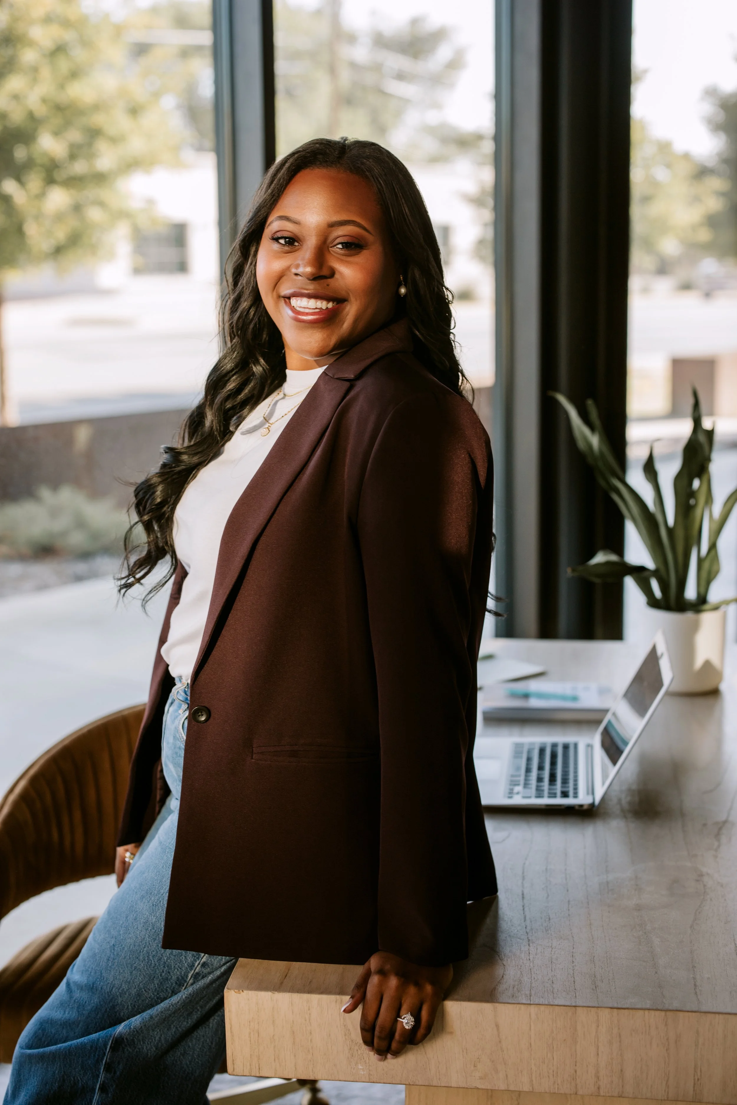 A woman with long, dark, wavy hair smiling and standing in an office, leaning on a wooden desk with a laptop and a potted plant.