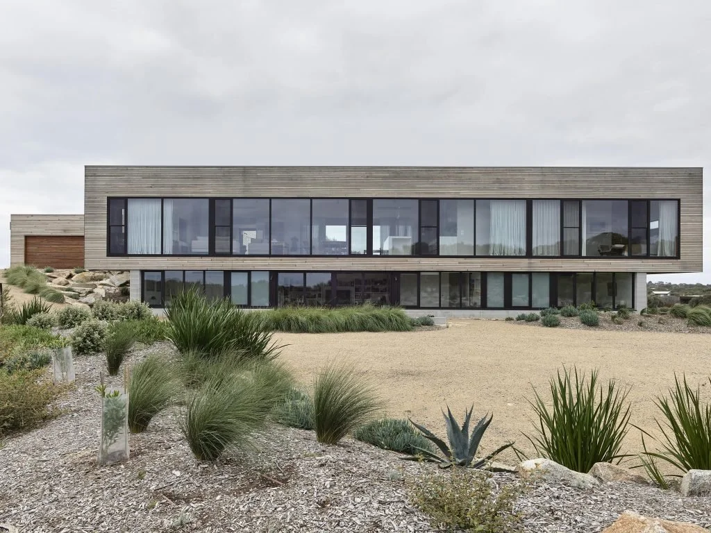 Modern two-story house with large glass windows and wooden exterior, surrounded by desert landscaping with various shrubs and plants.