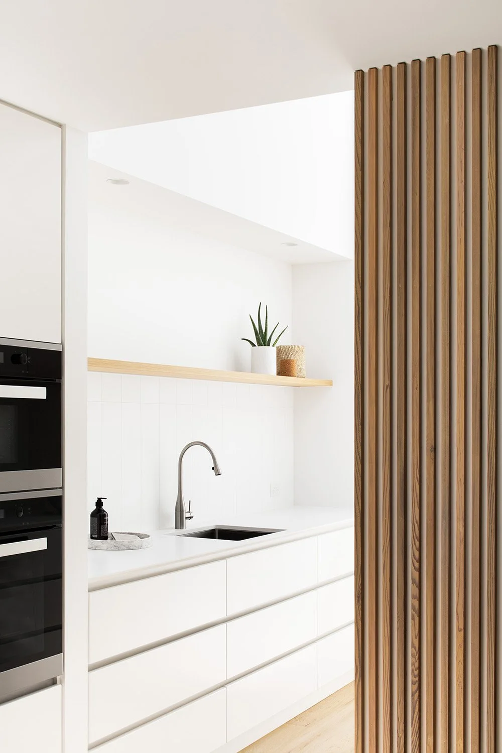 Modern minimalist kitchen with white cabinets, wooden accents, a black sink, and potted plants.