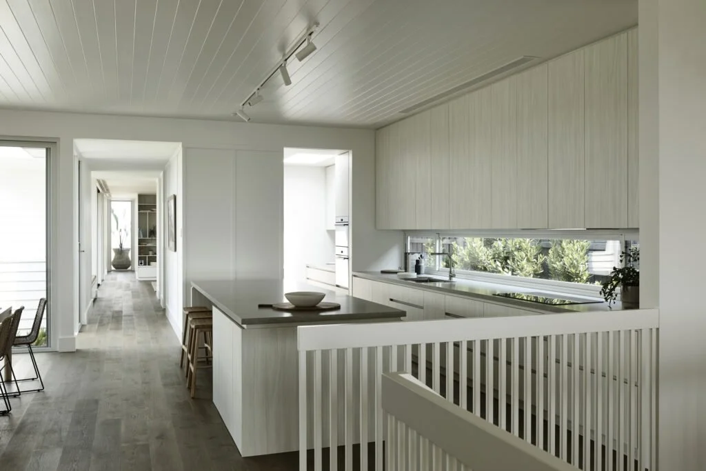 Modern, white kitchen with a large window, island with stools, and a hallway leading to other rooms.