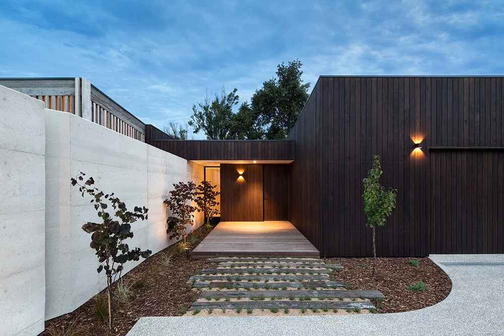Modern house exterior with dark wooden and white concrete walls, small plants, and ambient lighting at dusk.