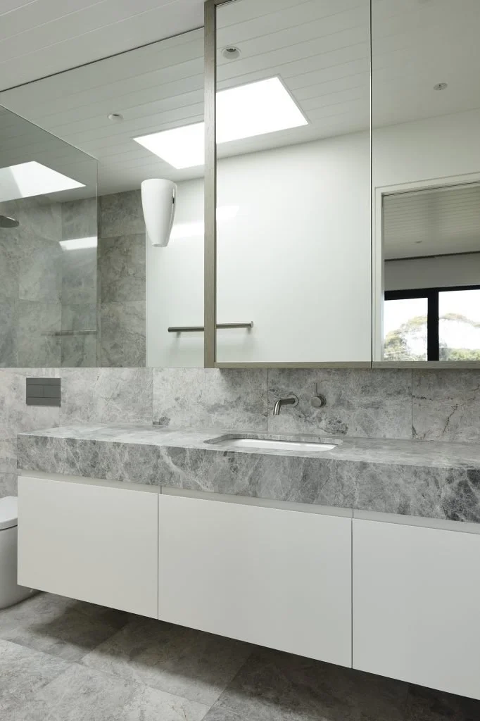 Modern bathroom with gray marble countertop and backsplash, white cabinets, a mirror, and a window showing trees outside.