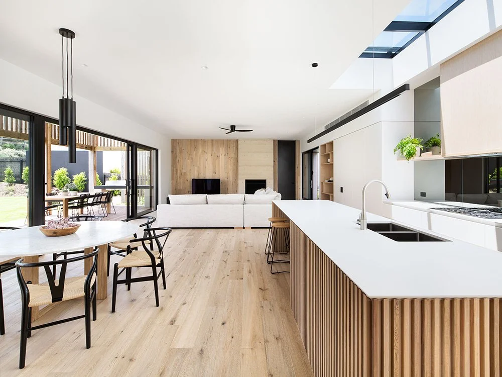 Open-concept living room and kitchen with modern, minimalist decor, wooden accents, large windows, and outdoor dining area.