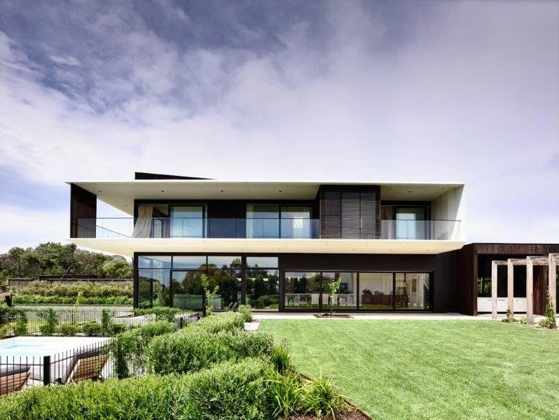 Modern two-story house with glass walls, large balcony, and landscaped yard