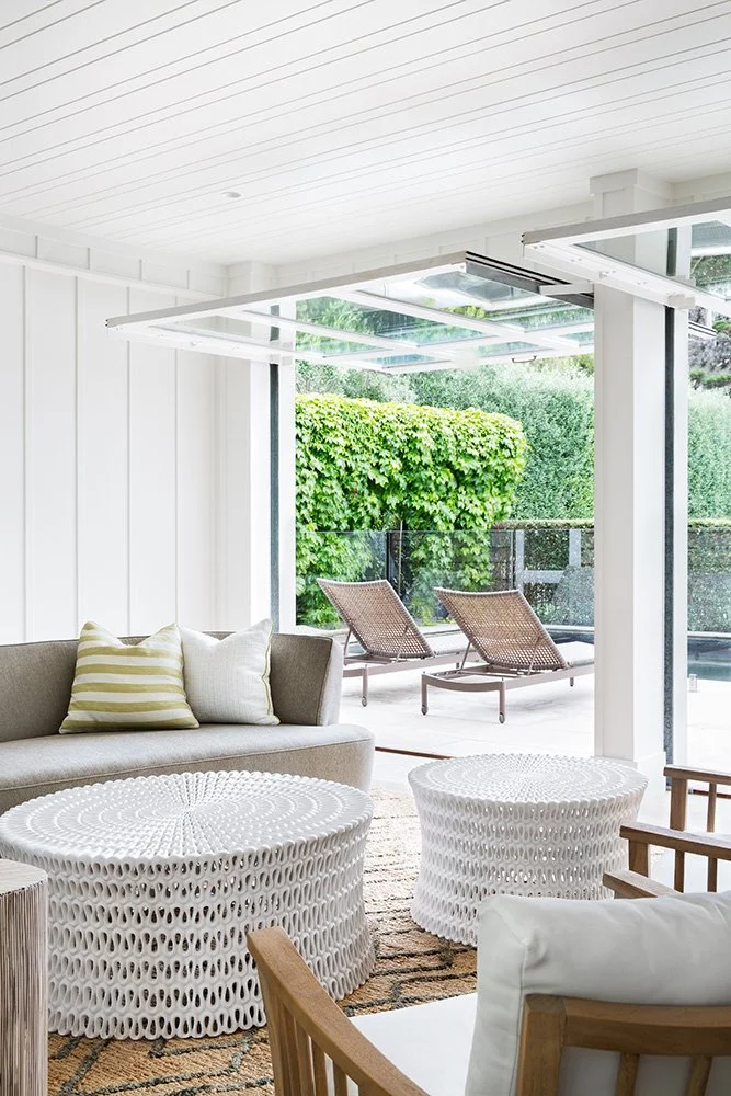 Living room with white wicker coffee tables, a beige sofa with striped and white pillows, a wooden armchair, and a view of outdoor patio with two lounge chairs and lush green bushes.