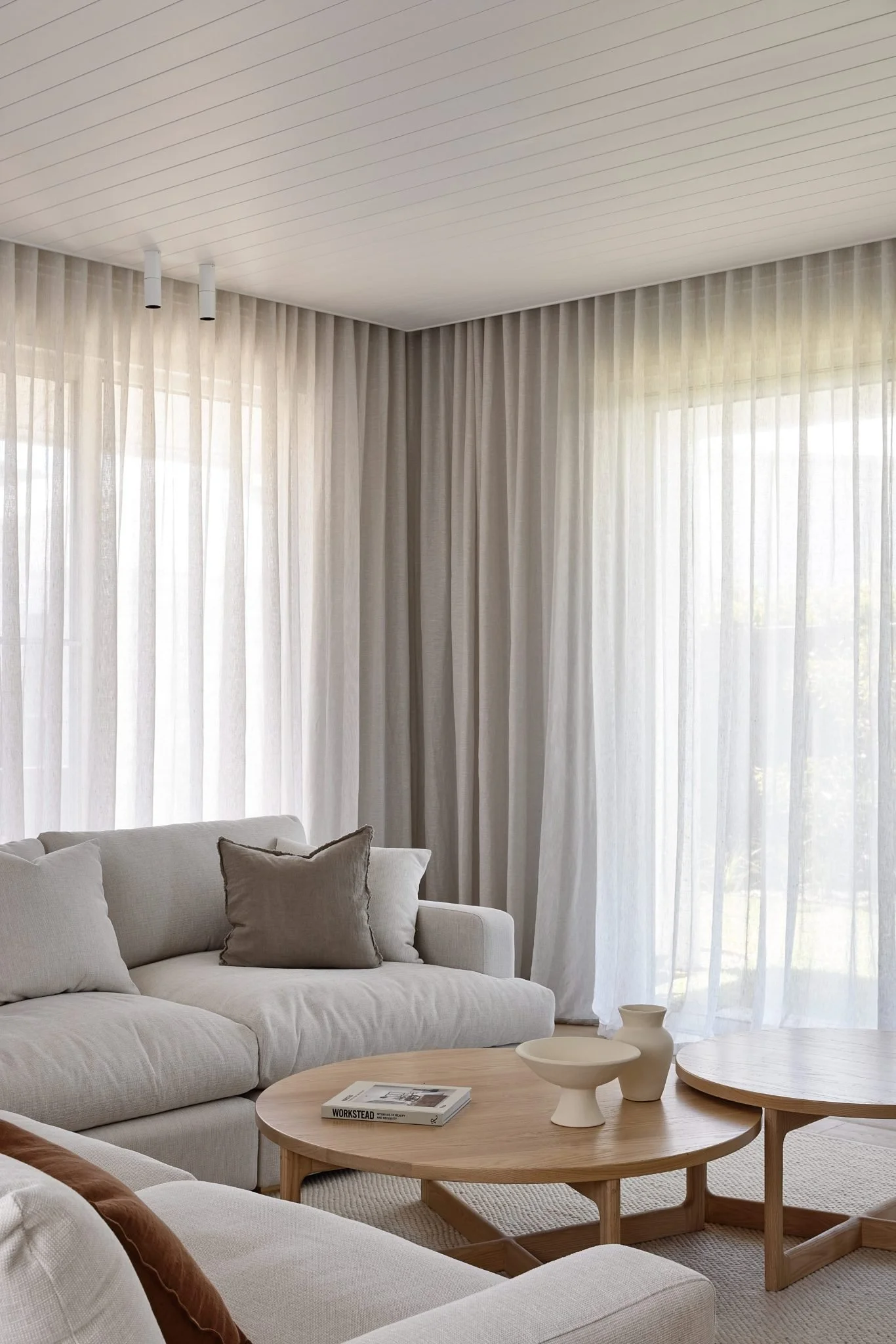 A bright living room with sheer white curtains, a cream sofa with matching pillows, and two round wooden coffee tables with ceramic vases and a magazine.