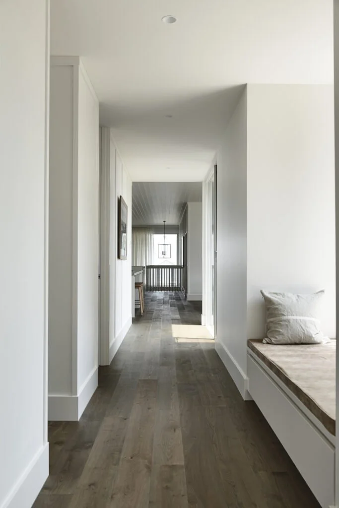 A bright, modern hallway with white walls, hardwood flooring, and a built-in bench with a pillow on the right. At the end of the hall, there is a room with a window, sheer curtains, and a hanging light fixture.