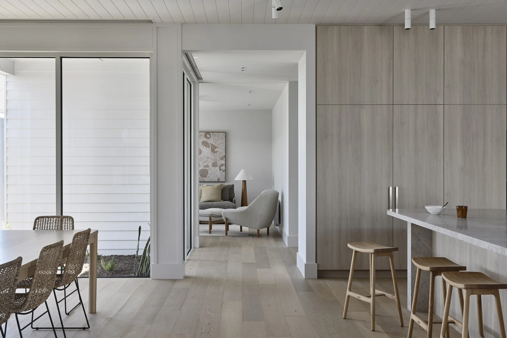 Modern minimalist living and dining area with light wood flooring, white walls, and natural wood furniture. The dining area has a table with woven chairs, and a small garden with plants is visible outside through sliding glass doors. The kitchen feat