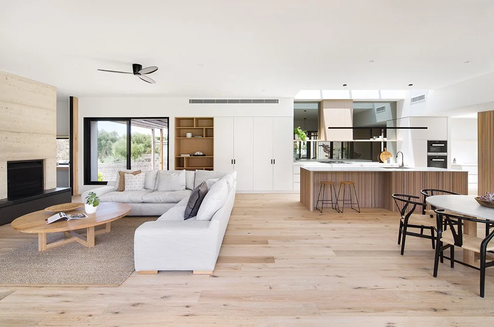 Open-concept living room and kitchen in a modern home with white walls, light wood flooring, a white sofa, a round wooden coffee table, a built-in fireplace, large windows, and a kitchen island with bar stools.