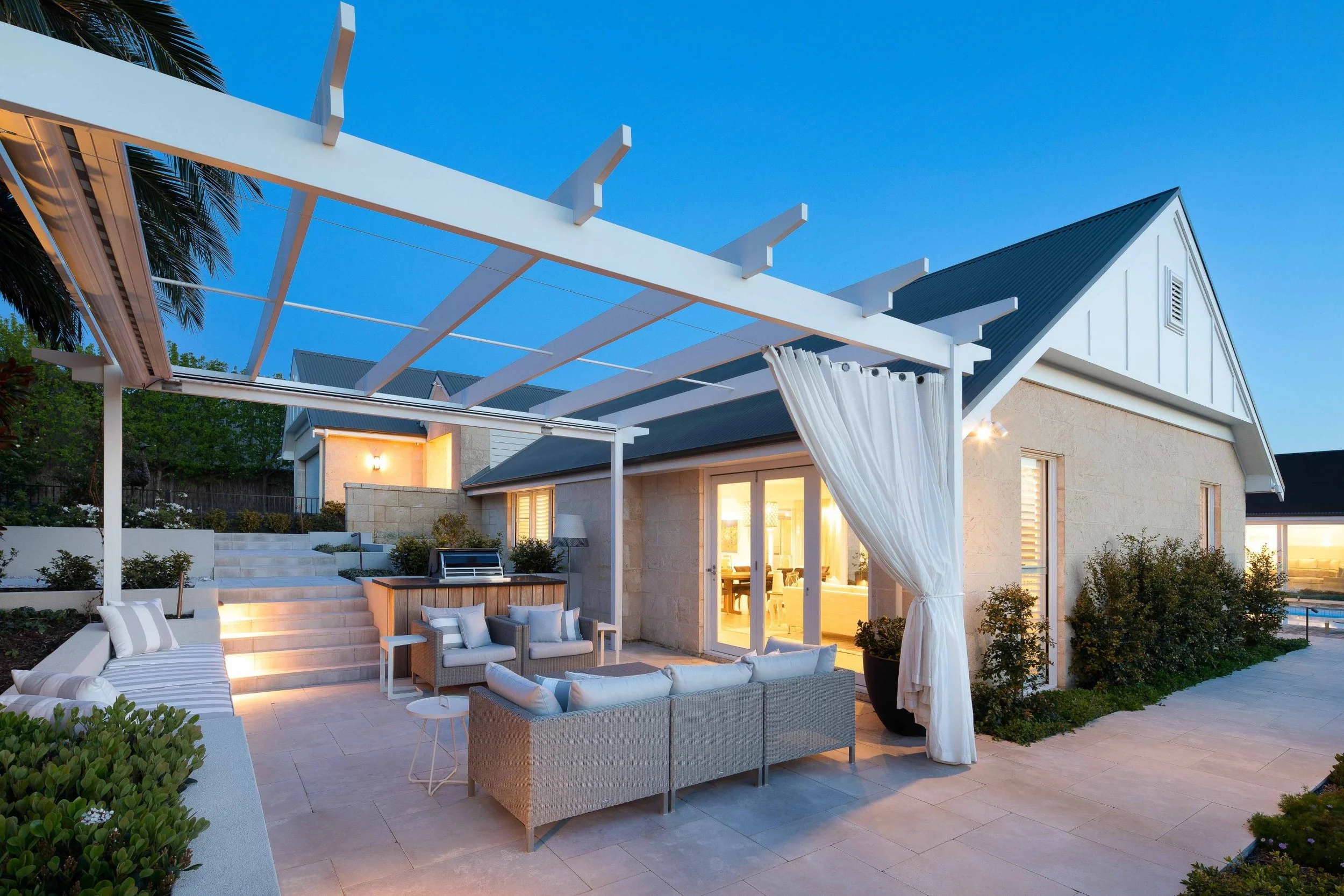Modern backyard patio with outdoor furniture, a pergola, and house with sliding glass doors, during evening with interior lights on.