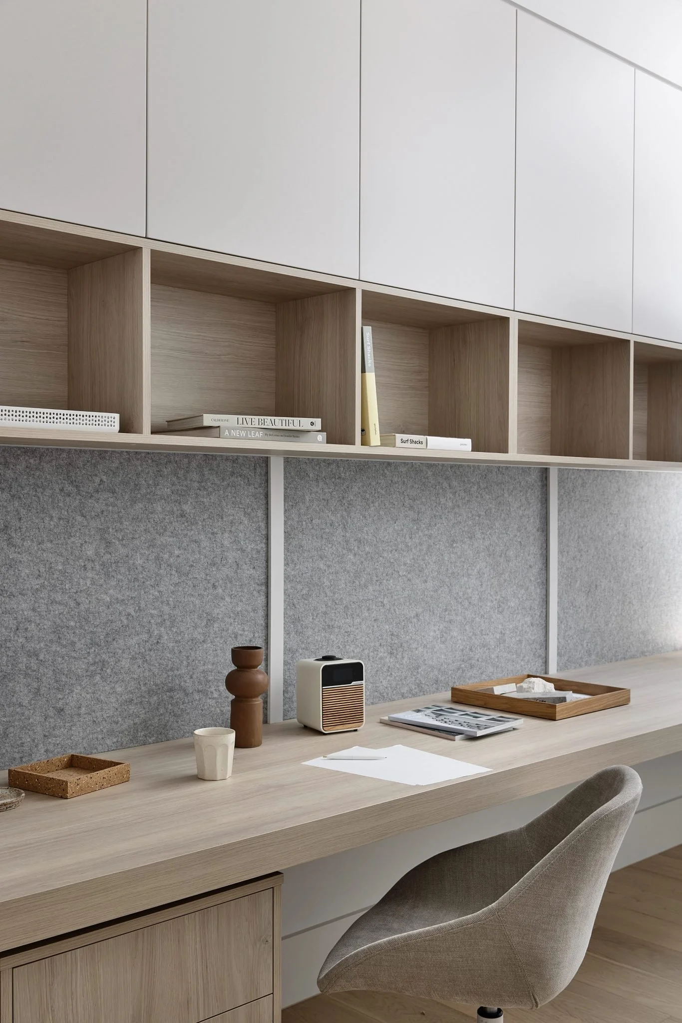 Modern workspace with a light wood desk, beige upholstered chair, gray fabric-covered panel, and upper wooden storage shelves with books and decor.