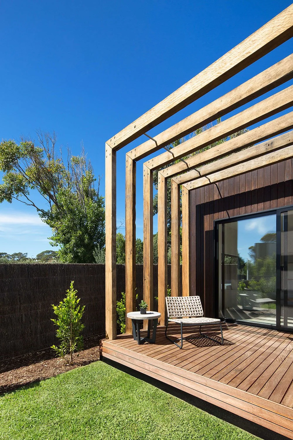 Backyard patio with wooden deck, minimalist bench, small plant, and wooden framing for a pergola on a sunny day with clear blue sky.