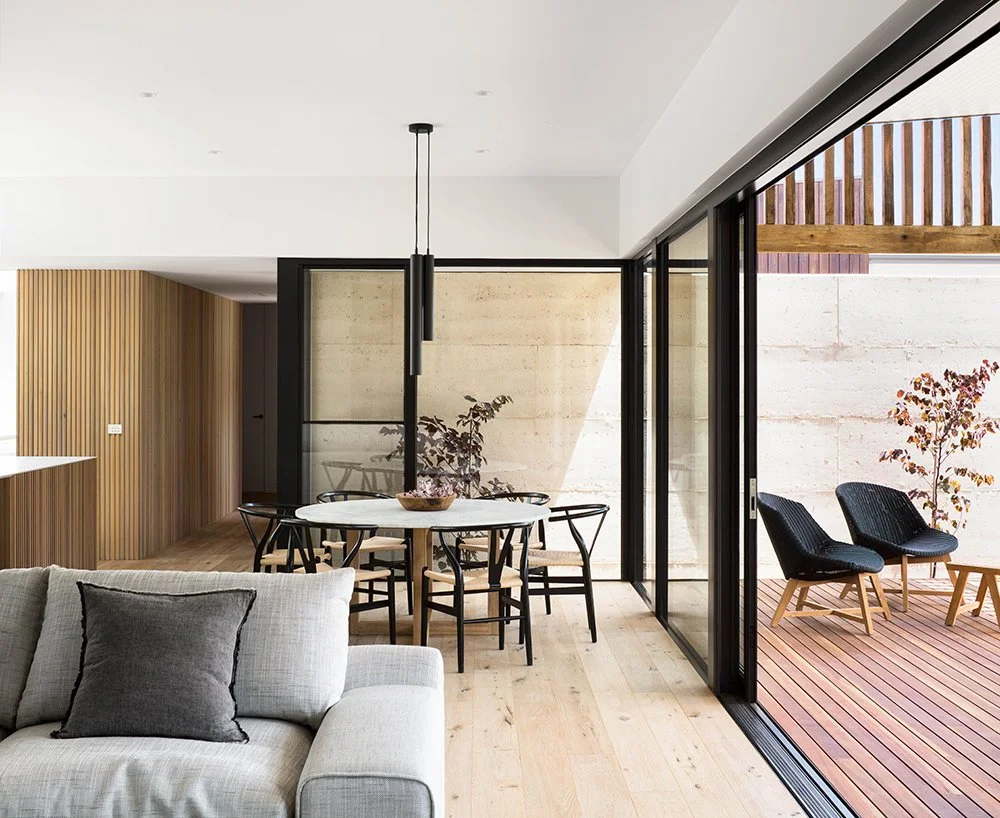 Modern living room with an open floor plan, featuring a white sofa with gray pillows, a round dining table with black chairs, a sliding glass door leading to an outdoor deck with two black chairs and a small tree.