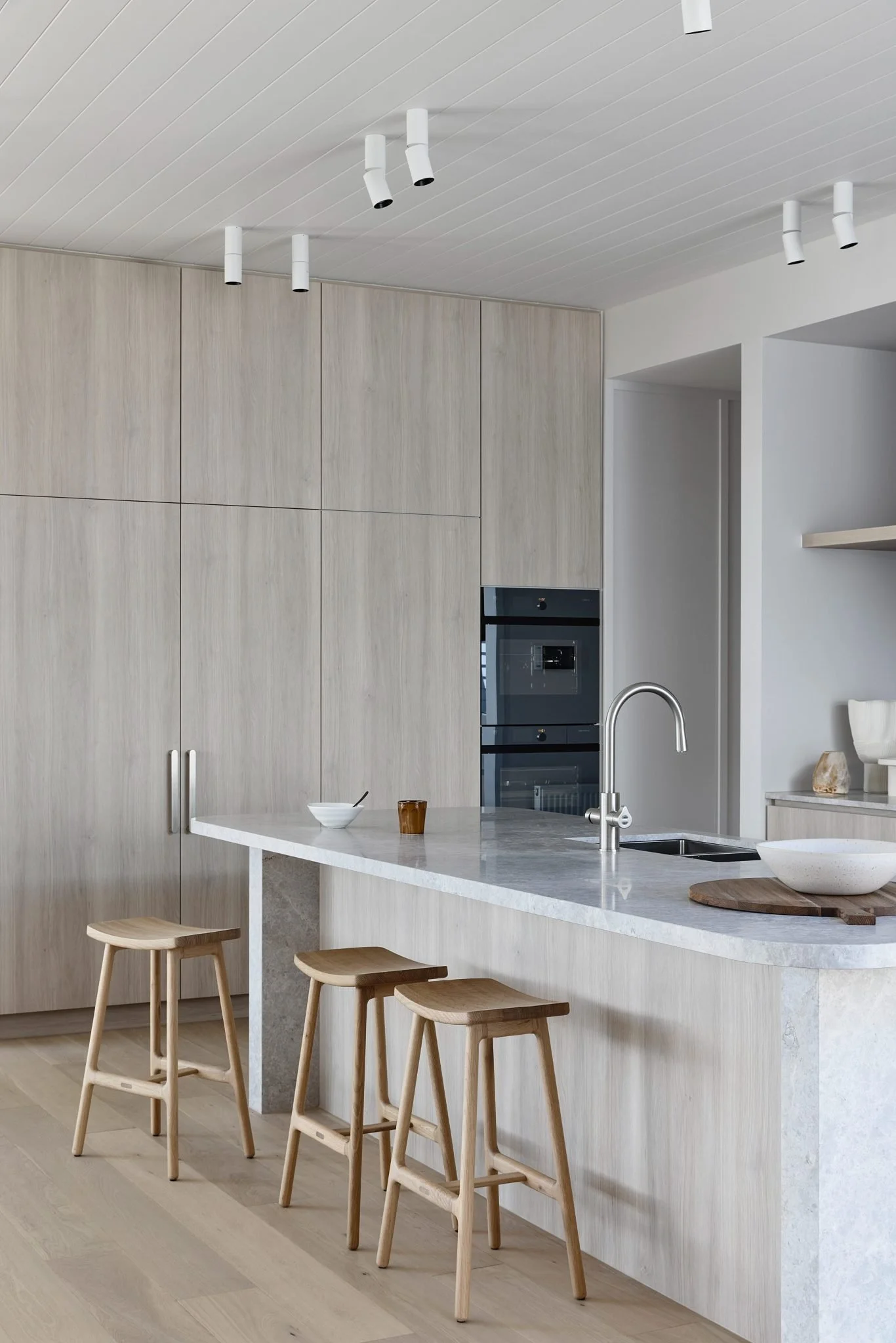 Modern kitchen with light wood cabinetry, a white marble island and three wooden barstools, stainless steel sink, and built-in oven and microwave.