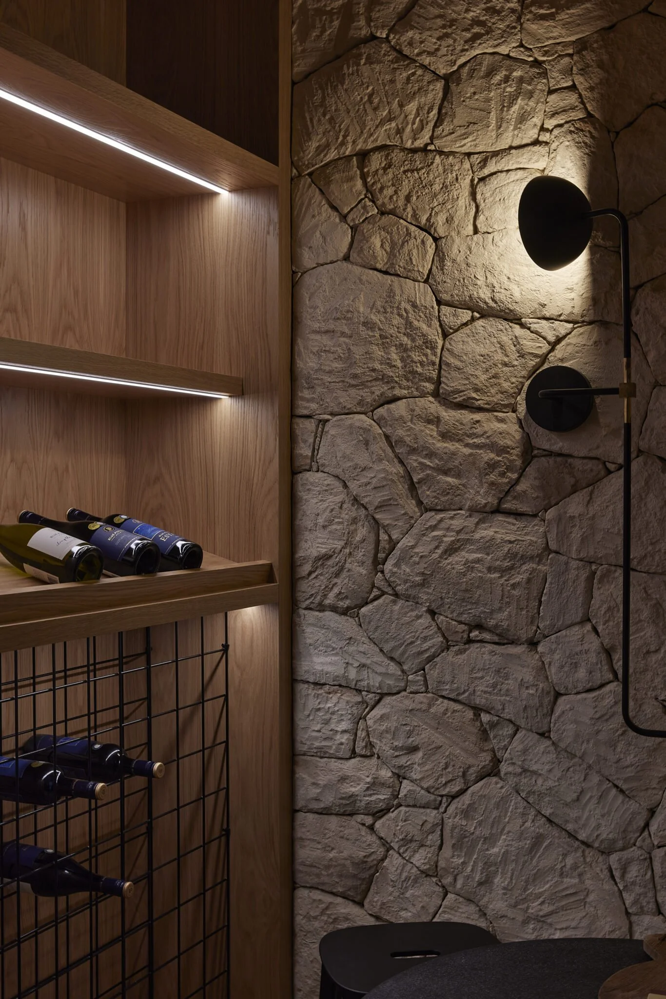 A cozy wine storage area with a stone wall, wooden shelves, and a black wall-mounted lamp.
