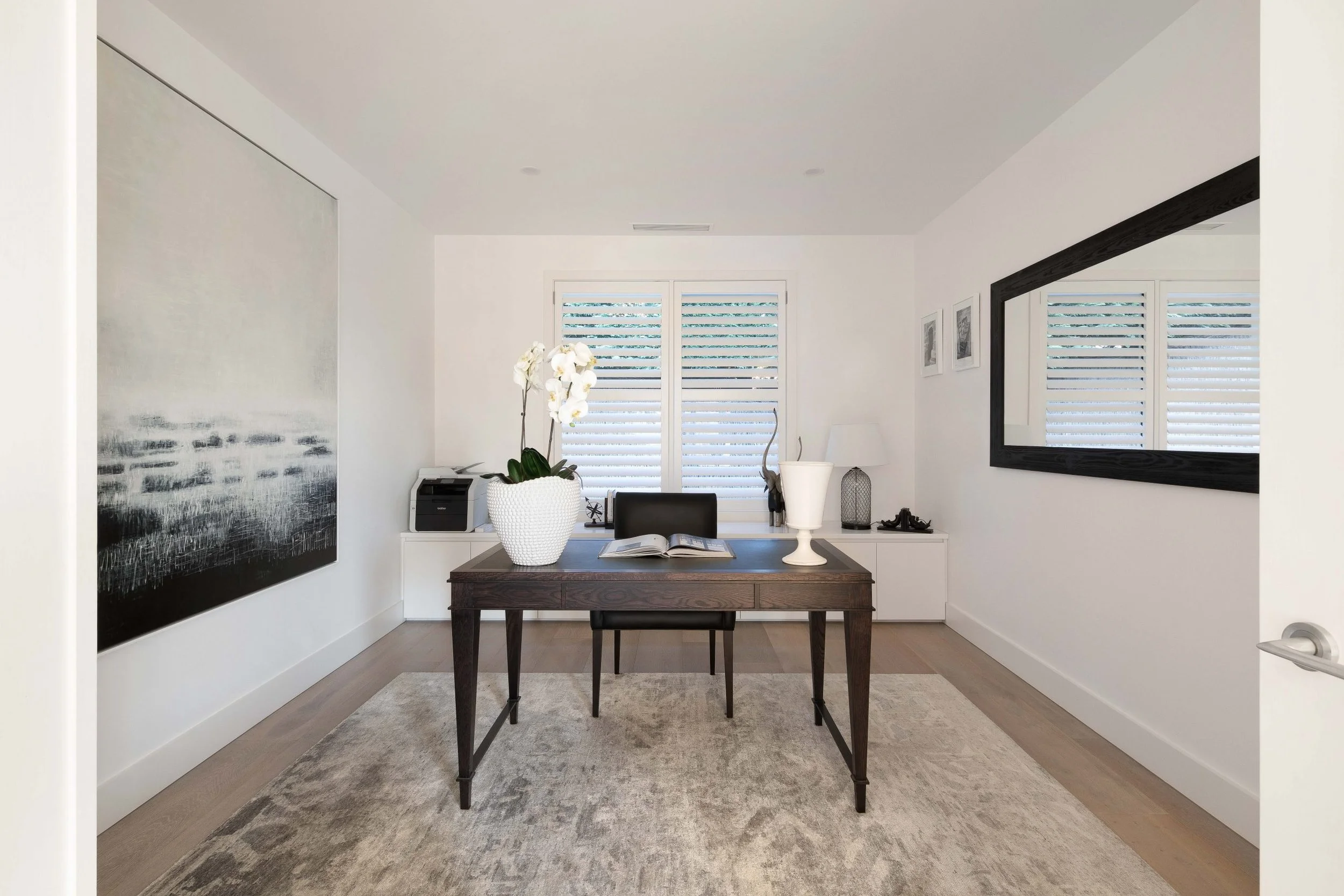 Modern home office with a dark wooden desk, black chair, white textured vase with orchids, open book, and large mirror on the right wall, white walls, and a window with blue and white blinds.