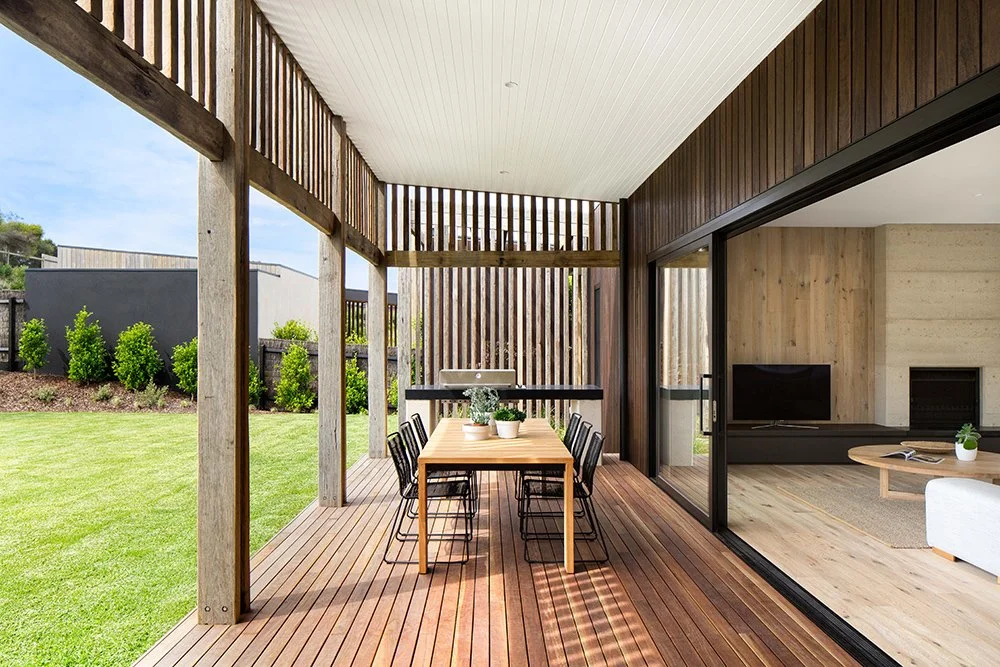 Outdoor patio with wooden deck, dining table with six chairs, surrounding grassy yard, and view into modern living room with fireplace and television.