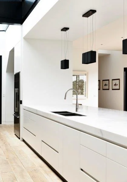 Modern minimalist kitchen with white cabinetry, a white countertop, a sink with a curved faucet, and black pendant lights hanging from the ceiling.