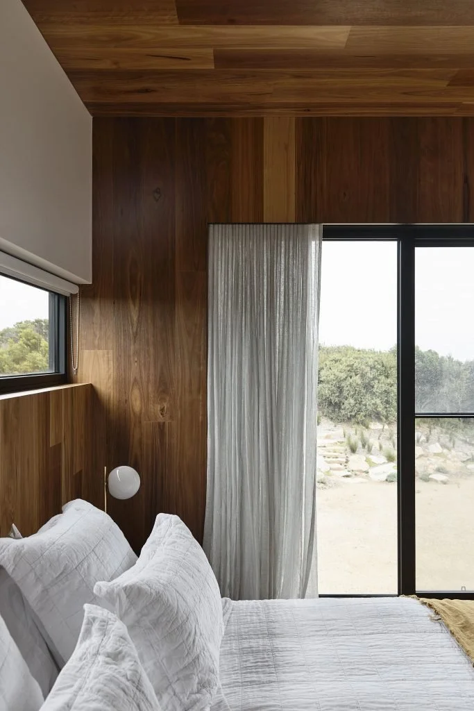 A bedroom with white bedding, wooden walls, a window with a roller shade, and sliding glass doors with sheer curtains leading outside.