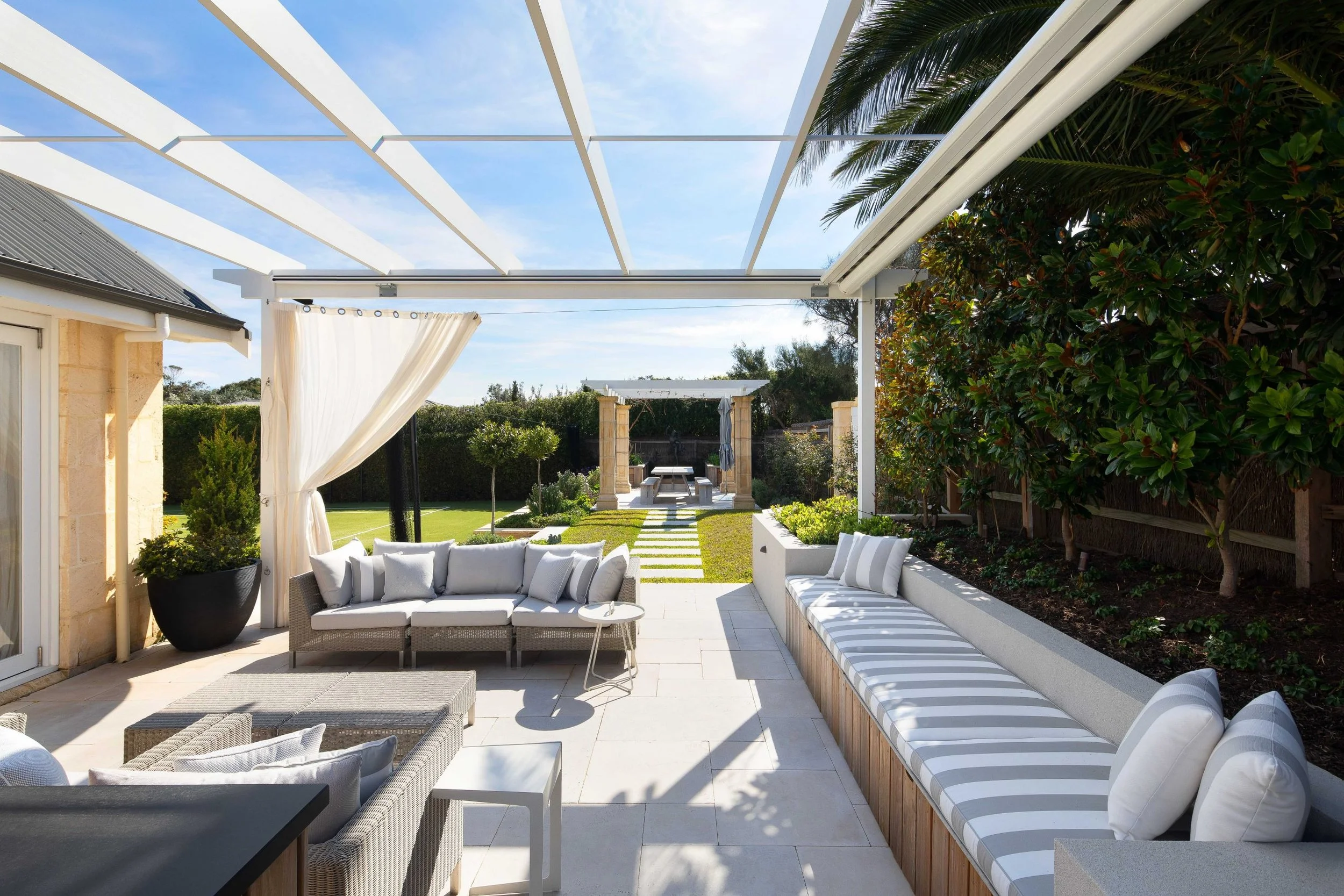 Outdoor patio with white cushioned seating, striped cushions, and a small table, overlooking a well-maintained garden and a pergola with a hot tub in the background.
