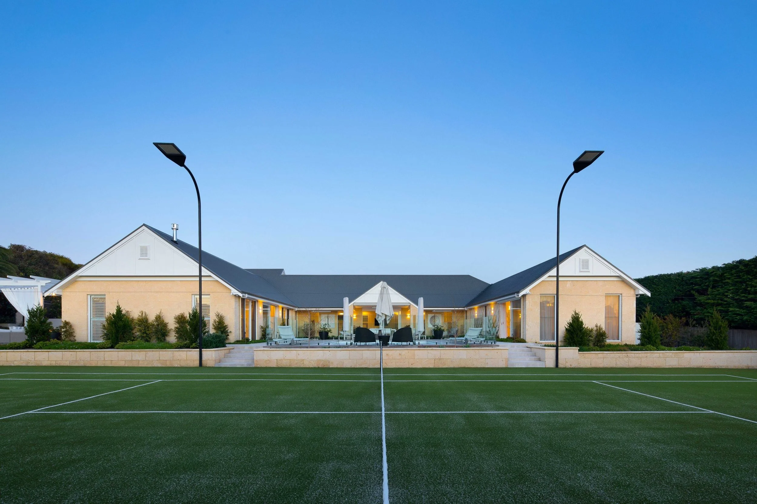 A modern house in the background with a well-maintained green tennis or sports court in the foreground, illuminated by two tall streetlights, during dusk or early evening.